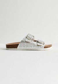 Silverfärgade platta sandaler med laserskurna mönster, dubbla spännen och en strukturerad korksula. Vit gummisula med zigzagkant.