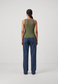 Olivengrønn ermeløs topp med blonde detalj på ryggen, kombinert med høytlivte blå vide bukser. Stoffet ser mykt og kroppsnært ut.