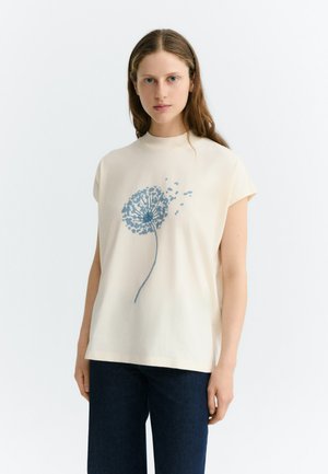 Jonge vrouw met lang bruin haar draagt een crème mouwloze top met een blauwe paardenbloemprint, gecombineerd met donkerblauwe broek.