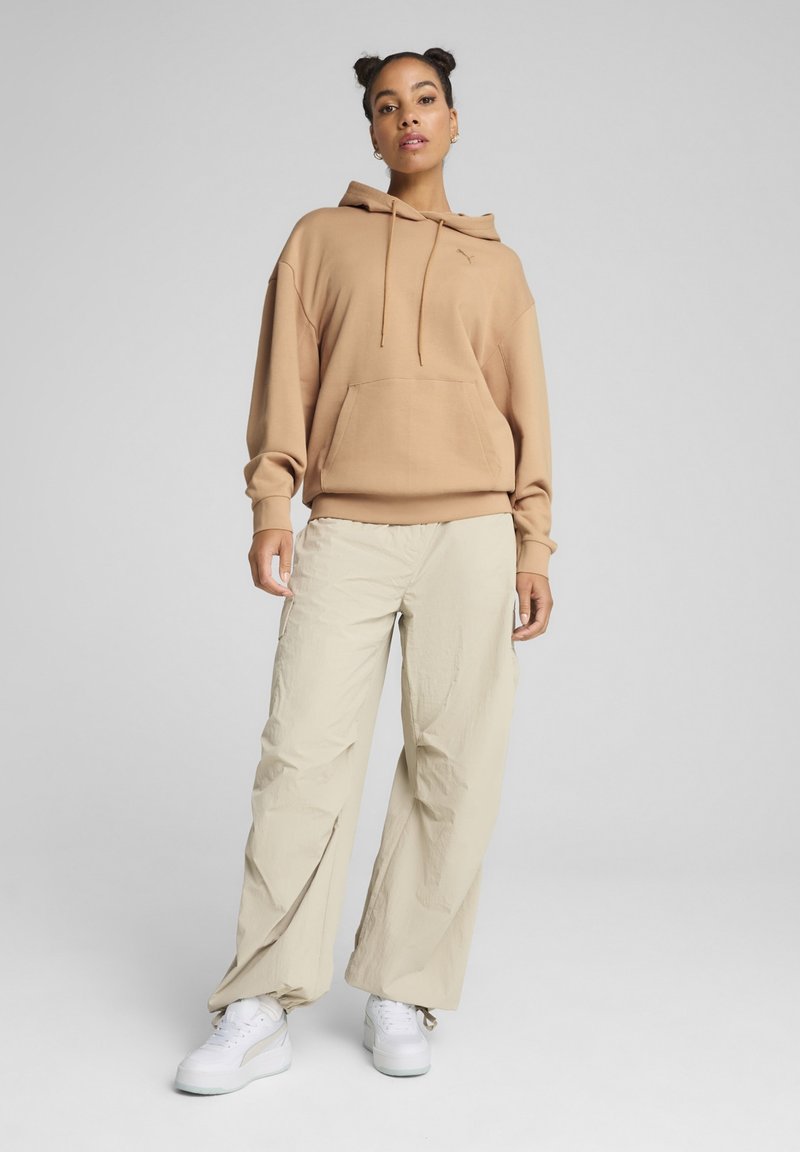 Felpa con cappuccio in cotone marrone chiaro con tasca frontale, abbinata a pantaloni beige larghi e sneakers bianche, caratterizzata da un design semplice.