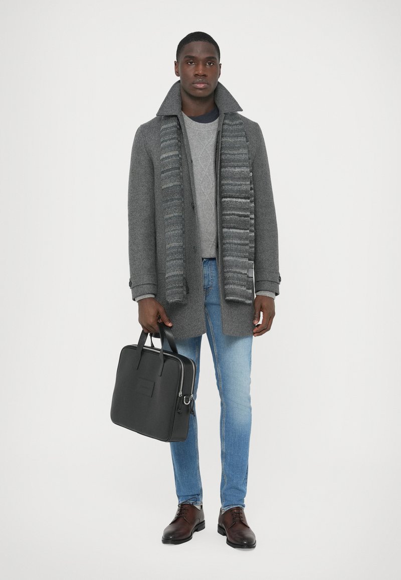 Cappotto di lana grigio con colletto alto, sciarpa grigia a righe, maglione grigio chiaro, jeans blu e scarpe di pelle marrone, con una borsa nera in mano.