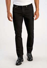 Jeans de mezclilla negra con corte ajustado. Presentan herrajes de cobre y un diseño clásico de cinco bolsillos. Combinados con unos mocasines negros brillantes.