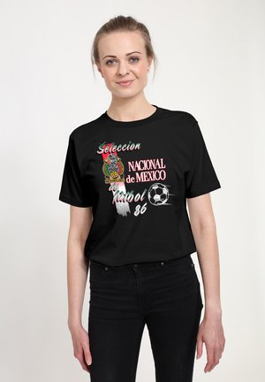 Kvinde med lyst hår i en sort T-shirt med Mexico landsholdets fodboldlogo, tekst "Seleccion Nacional de Mexico futbol 86" og en grafik af en fodbold.