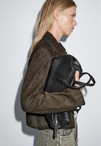 Black leather tote bag with textured finish held against an olive suede jacket, featuring zippers and sturdy handles. Classic casual design.
