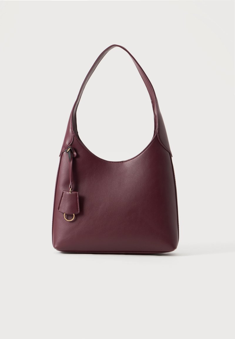 Sac à main en cuir bordeaux foncé avec une bandoulière simple et une petite étiquette décorative avec un anneau doré sur un fond clair uni.