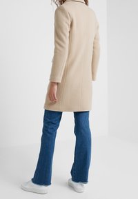 Beige Wollmischungsjacke mit Kragen, langen Ärmeln, taillierter Passform und Rückennahtdetails, kombiniert mit ausgestellten blauen Jeans und weißen Sneakers.