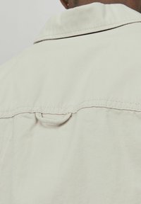 Veste beige clair au fit décontracté, dotée d'un col, de détails de couture et d'une boucle à l'arrière pour l'accrocher. Texture lisse.