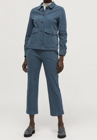 Modrý dvědílný outfit z manšestru, obsahující bundu s límečkem a knoflíkovým uzávěrem a dvěma předními kapsami, spárovaný s zkrácenými kalhotami a černými botami.