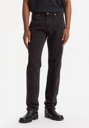 Persoon in zwarte rechte jeans, zwarte leren laarzen en een zwart topje, staande tegen een witte achtergrond.