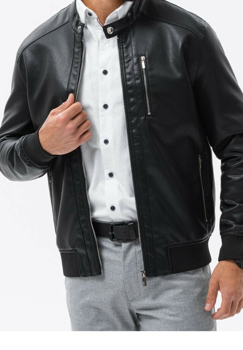 Zwarte leren jas met een rits aan de voorkant en een opstaande kraag. Voorzien van zijzakken en geribbelde manchetten. Draagt over een witte shirt.