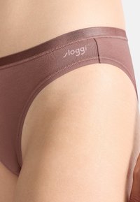 Braune Bikini-Hose aus weichem Stoff mit glatter Textur und einem Logo auf dem elastischen Bund. Für einen bequemen Sitz geschnitten.