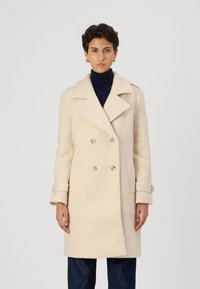 Krēmkrāsas dubultījuma mētelis ar lieliem atlokiem, teksturētu audumu, zelta toņa pogām un sānu kabatām, savienots ar tumšu turtleneckreklu.