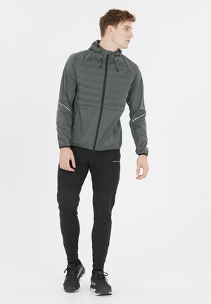 Veste d'extérieur grise avec fermeture éclair à l'avant, capuche et détail matelassé sur la poitrine, associée à un pantalon noir ajusté et des chaussures de course sombres.