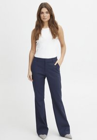 Navy tailored trousers with a smooth texture, featuring front pockets and a slim fit. Paired with a white sleeveless top and gray heels.