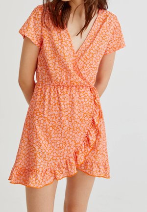 Frau trägt ein orangefarbenes Wickelkleid mit kleinem rosa Blumenmuster und Rüschensaum, steht mit den Händen auf dem Rücken.
