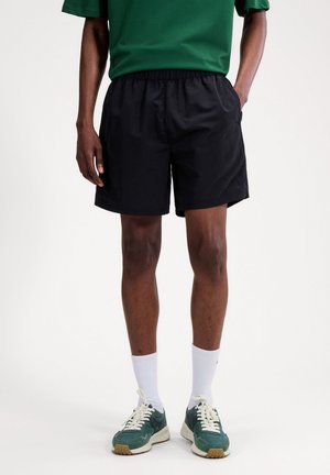 Hombre con pantalones cortos negros de cintura elástica, zapatillas verdes, calcetines blancos hasta el tobillo y una camiseta verde de manga corta frente a un fondo liso.