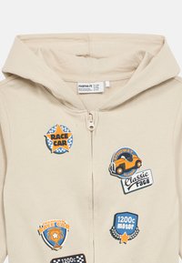 Beige hettejakke med glidelås og hette. Har fargerike, racing-inspirerte merker: «Race Car», «Classic Race» og «1200c motor». Myk tekstur.