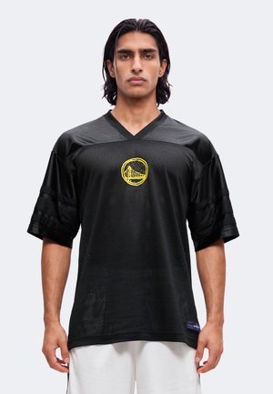 Homme aux cheveux noirs mi-longs, portant un maillot de sport ample en maille noire avec un logo de pont jaune et un short blanc sur un fond uni.