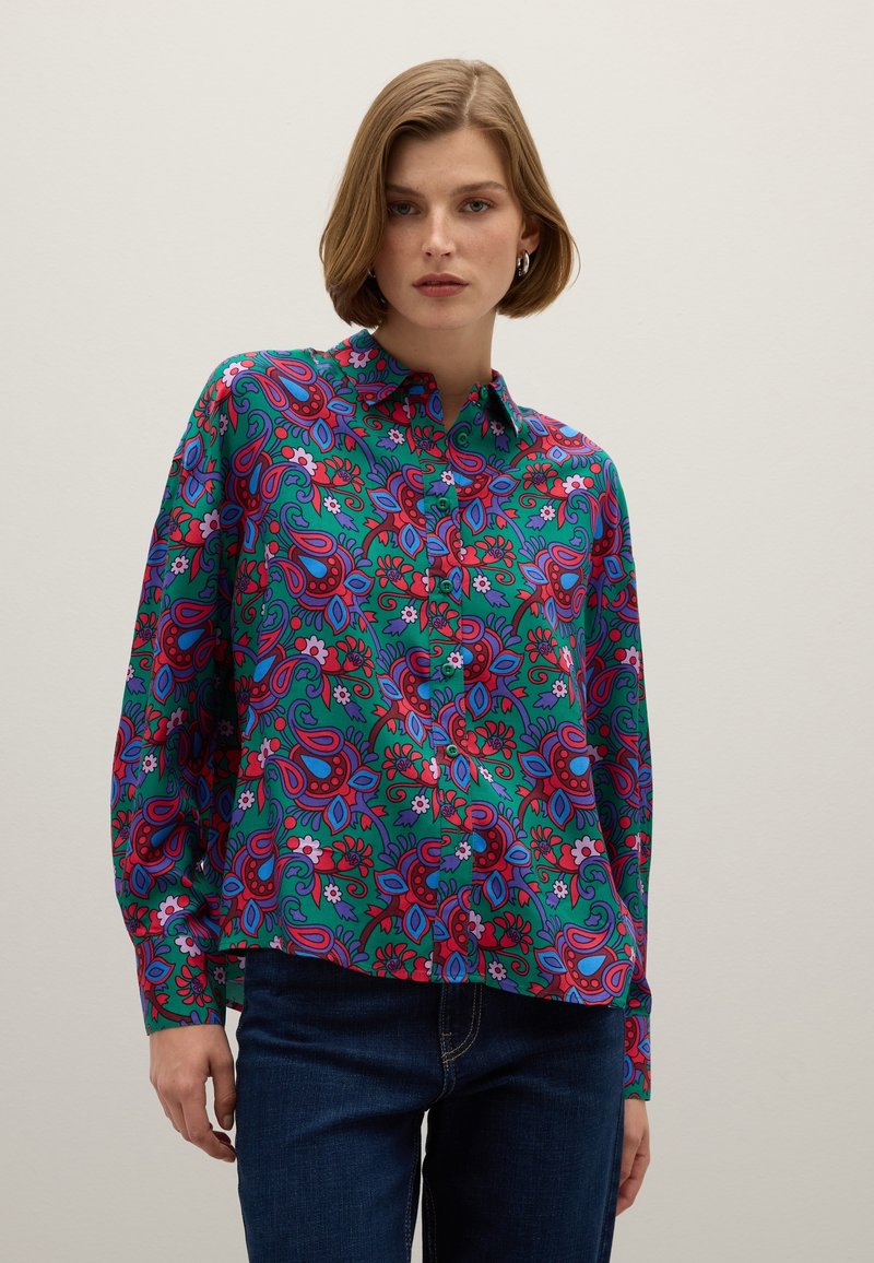 Donna che indossa una camicia a maniche lunghe con un vivace motivo floreale in verde, rosso e viola, abbinata a dei jeans blu, su uno sfondo chiaro e semplice.