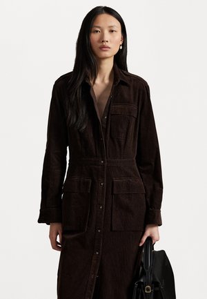 Bruine corduroy jurk met lange mouwen, knoopsluiting aan de voorkant, twee borstzakken en een getailleerde pasvorm, gecombineerd met een zwarte handtas.