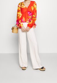 Vloeiende, rode bloemenblouse met lange mouwen en een V-hals. Gecombineerd met witte, wijde broek en plateau sandalen. Inclusief een kleine geweven tas.