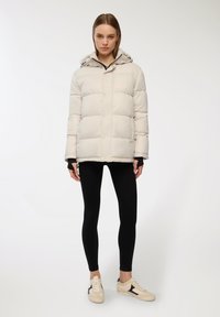 Giacca puffer beige chiaro con cappuccio, caratterizzata da cuciture orizzontali, bottoni a pressione e tasche laterali, abbinata a leggings neri e sneakers.