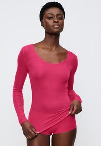 Langärmliger Body in einfarbigem Pink mit geripptem Textil. Rundhalsausschnitt und figurbetonter Schnitt. Das Modell hält den Stoff an der Taille und zeigt die Form des Kleidungsstücks.