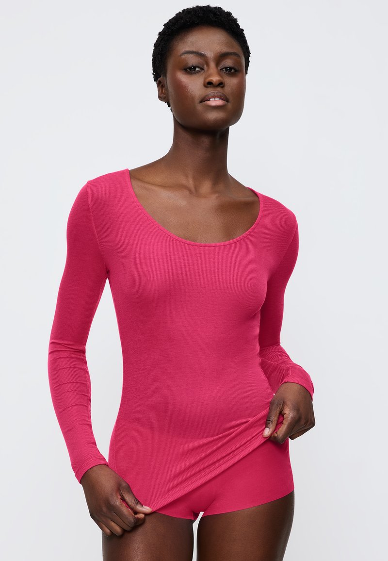 Langärmliger Body in einfarbigem Pink mit geripptem Textil. Rundhalsausschnitt und figurbetonter Schnitt. Das Modell hält den Stoff an der Taille und zeigt die Form des Kleidungsstücks.