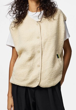 Persoon draagt een mouwloos beige sherpa-vestje met knopen over een wit T-shirt en zwarte broek, met zichtbaar krullend donker haar.