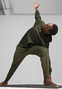 Olivegrünes Sportoutfit bestehend aus einem Kapuzenshirt und weiten Hosen, mit einer weichen Textur. Die Teilnehmerin befindet sich in einer Yoga-Pose auf einer Matte.