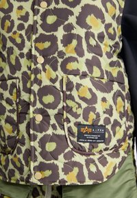 Strømlinjeformet quiltet vest med et grønt og brunt leopardmønster, gyldne knapper, to frontlommer og et merkeetikett.