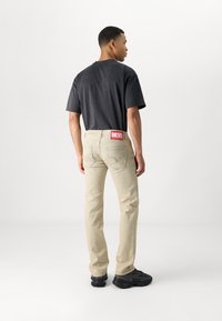 Jeans de mezclilla beige con un diseño ajustado, que cuentan con una etiqueta roja "DIESEL" en la parte trasera de la cinturilla y costuras en contraste. Combinados con una camiseta negra.