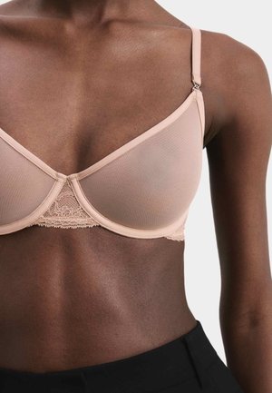 Eine dunkelhäutige Person trägt einen hautfarbenen Mesh-BH mit Spitzenverzierungen und schwarze Hosen, von der Hals- bis zur Taille abgebildet.