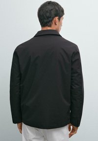 Man wearing a black long-sleeve jacket and white pants, standing facing away against a plain light background.