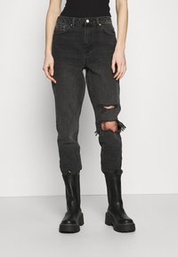 Jeans de mezclilla negros con desgastes y roturas en las rodillas, diseño de cintura alta y piernas ajustadas, combinados con botas negras hasta la rodilla.