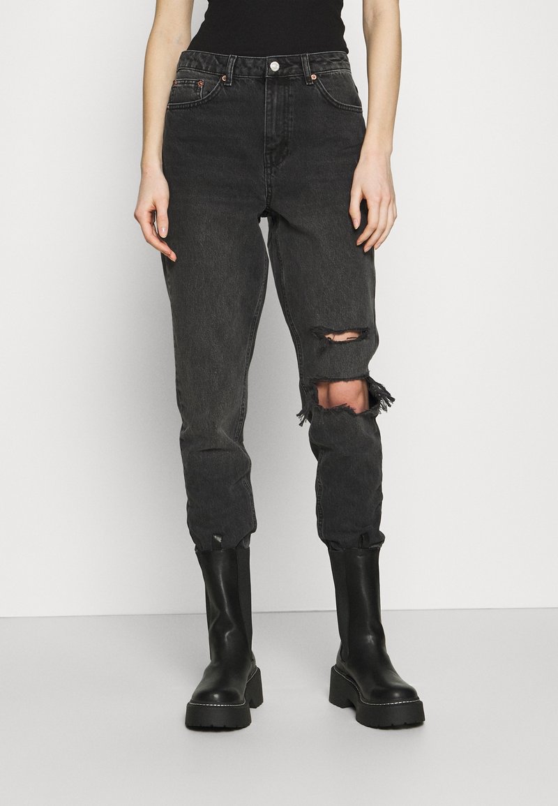 Jeans de mezclilla negros con desgastes y roturas en las rodillas, diseño de cintura alta y piernas ajustadas, combinados con botas negras hasta la rodilla.