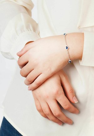 Bracciale in argento con catena, caratterizzato da gemme blu a forma di pera e rotonde, indossato su un polso con una camicetta di colore chiaro.