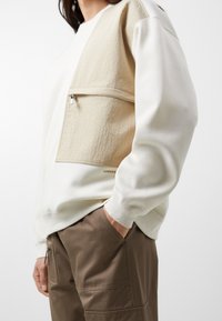 Sudadera crema con un panel texturizado beige, bolsillo con cremallera, puños acanalados y un pequeño detalle del logo cerca del dobladillo. Combinada con pantalones marrones.