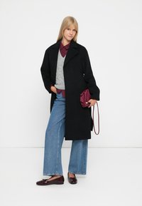 Cappotto di lana nera con colletto, indossato sopra un maglione grigio e una camicia bordeaux. Jeans a gamba larga e borsa bordeaux completano il look.