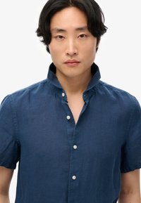 Jeune homme aux cheveux noirs mi-longs portant une chemise bleu marine à manches courtes boutonnée avec le col relevé.