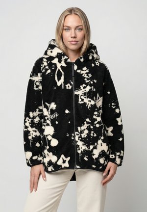 Frau trägt eine schwarze Fleecejacke mit weißen abstrakten Blumenmustern und Kapuze, kombiniert mit hellen Hosen, stehend vor grauem Hintergrund.