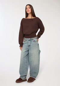 Młoda kobieta z prostymi brązowymi włosami nosi luźny brązowy sweter, szerokie jasnoniebieskie dżinsy i brązowe buty wsuwane, stojąc na białym tle.