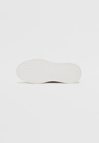 Black sneaker with a white rubber sole, featuring a textured grip pattern and a logo embossed on the center. Smooth upper surface.