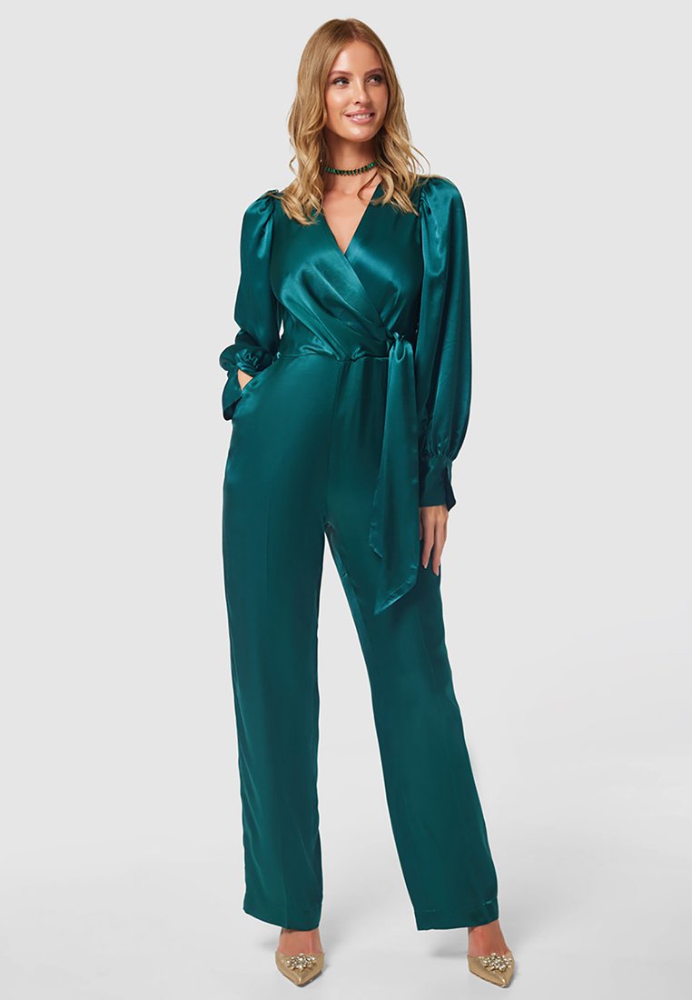 Tannengrüner Satin-Jumpsuit mit Wickel-Front, langen puffigen Ärmeln und gefaltetem Taillengürtel. Kombiniert mit spitzen High Heels, die eine elegante Silhouette schaffen.