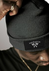Black knit beanie with vertical ribbed texture and a flat cuff. Features a rectangular black patch with the word "valhalla" and axe graphic.