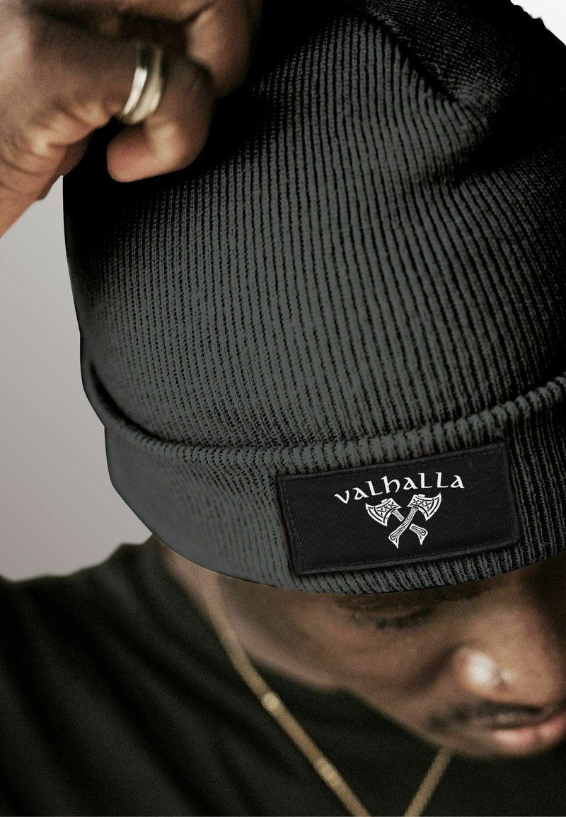 Black knit beanie with vertical ribbed texture and a flat cuff. Features a rectangular black patch with the word "valhalla" and axe graphic.