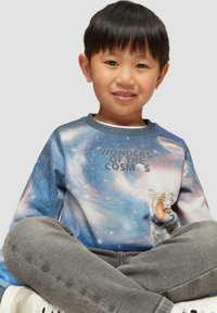 Langarmpullover met een kosmische print in blauwtinten, een tijgerafbeelding en de tekst "Wonderen van de Cosmos" in grijs. Grijze broek en witte schoenen.