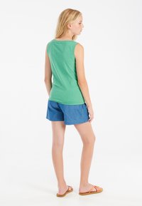 Top senza maniche verde abbinato a pantaloni shorts in denim blu. Il top ha un collo rotondo, mentre gli shorts sono di lunghezza al ginocchio. Sandali a infradito marroni indossati.