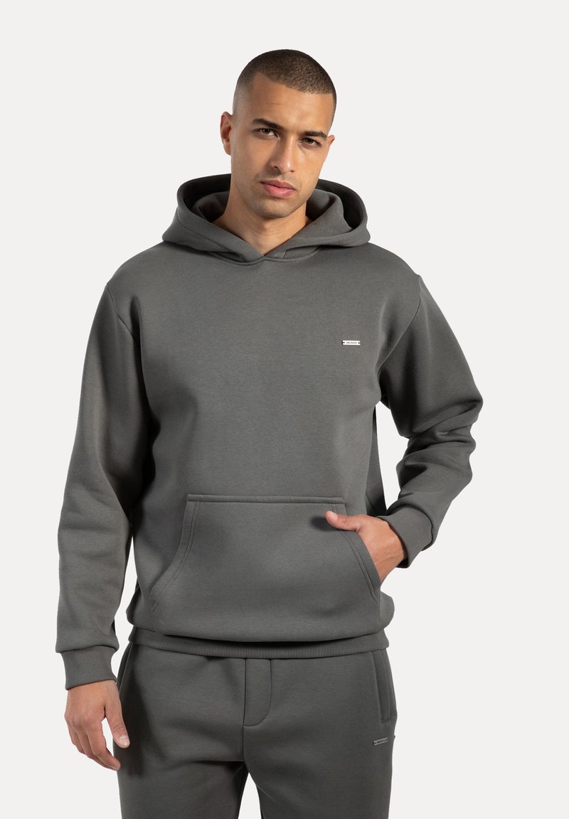 Sudadera con capucha gris de tejido suave, con un bolsillo frontal, puños de canalé y un pequeño emblema en el pecho. Textura lisa.