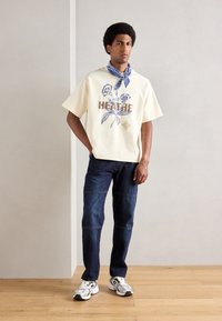Lys beige grafisk T-shirt med blomsterdesign og brun tekst. Paireret med mørkeblå jeans og hvide sneakers med grå accenter.